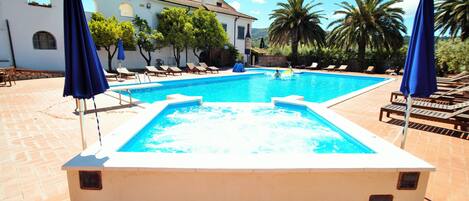 A rectangular swimming pool with a hot tub, surrounded by lounge chairs and umbrellas, set against a backdrop of a white building with arched windows and palm trees.