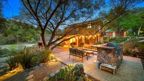 A patio area with outdoor furniture, a stone barbecue, and a view of a house in the background. - Luxury Riverfront Paradise Escape (Three Rivers)