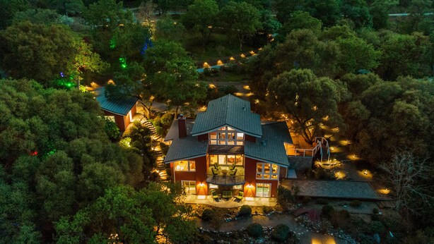 A large, two-story house surrounded by dense trees, with a visible patio area and outdoor lighting. - Luxury Riverfront Paradise Escape (Three Rivers)