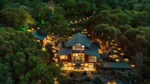 A large, two-story house surrounded by dense trees, with a visible patio area and outdoor lighting. - Luxury Riverfront Paradise Escape (Three Rivers)