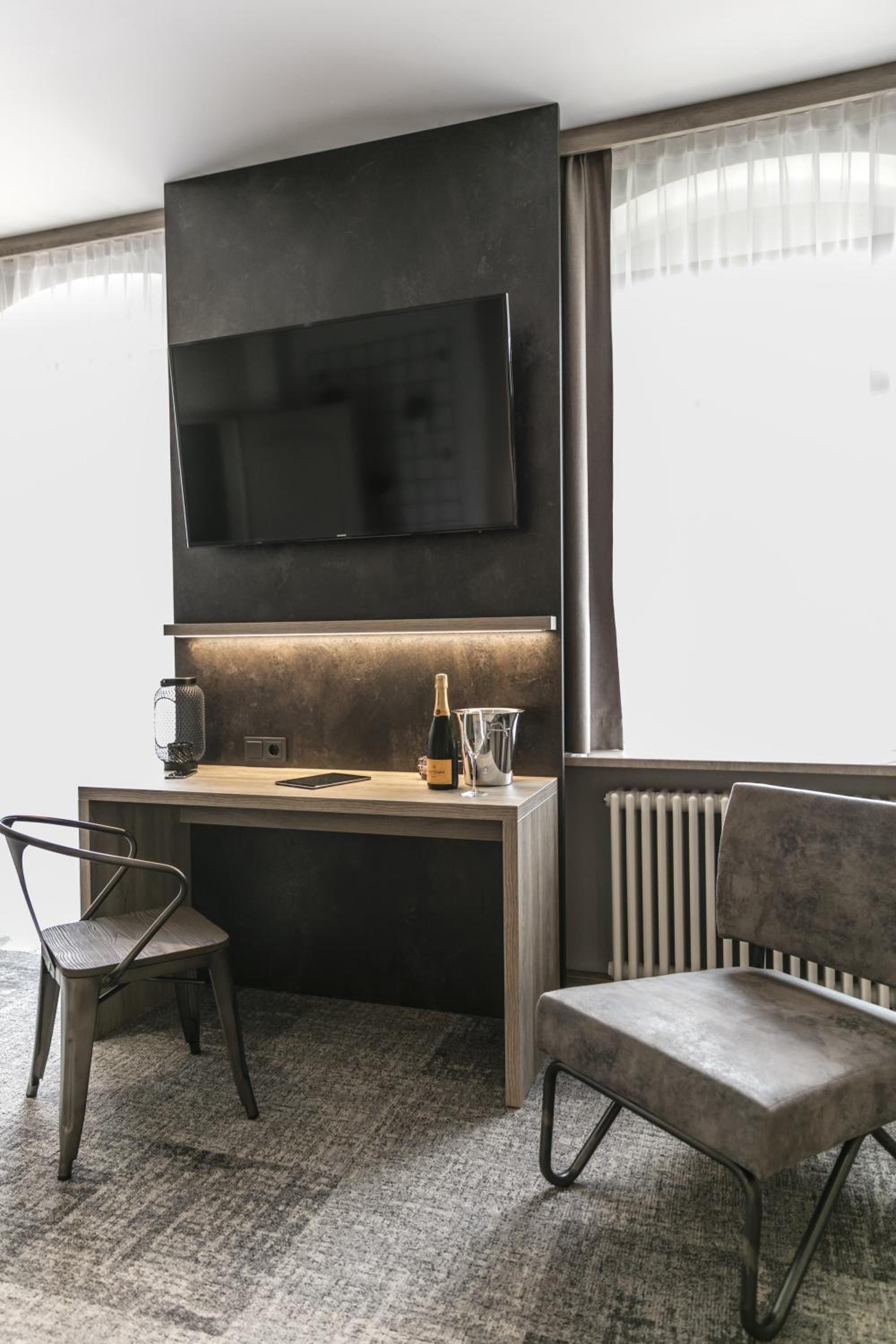 A modern hotel room with a flat-screen TV mounted on the wall, a wooden desk with a bottle of champagne, a glass, and a smartphone, two chairs, and a radiator.