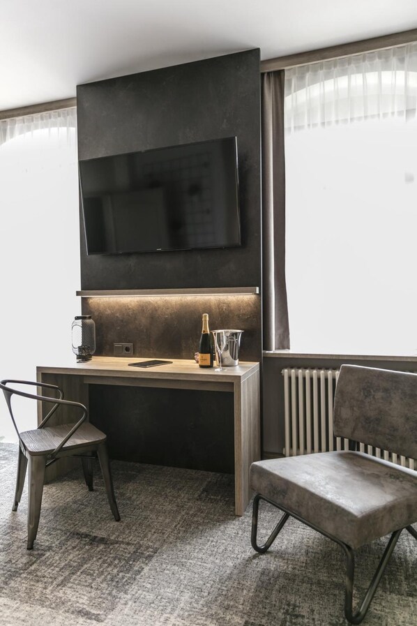Una habitación de hotel moderna con una televisión de pantalla plana montada en la pared, un escritorio de madera con una botella de champán, una copa y un teléfono inteligente, dos sillas y un radiador.