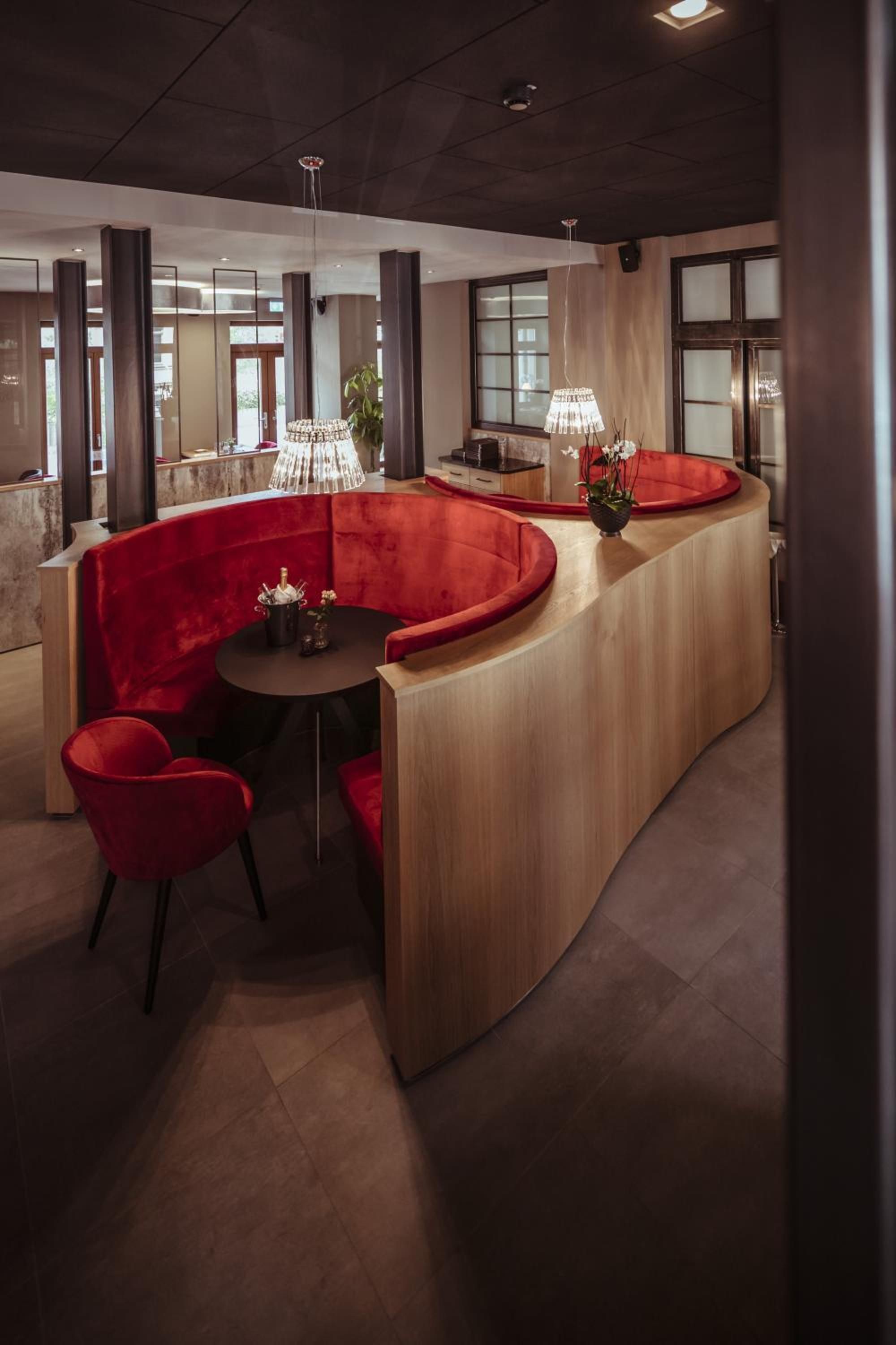 Un área de descanso moderna con un banco de madera curvo, sillas tapizadas en rojo y una mesa redonda.