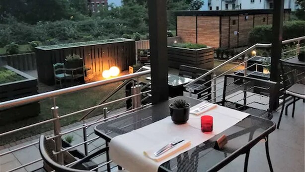 An outdoor dining area with a table, chairs, and a railing. There is a building with a brick facade in the background. - Alexxanders Restaurant & Bordinghouse (Chemnitz)