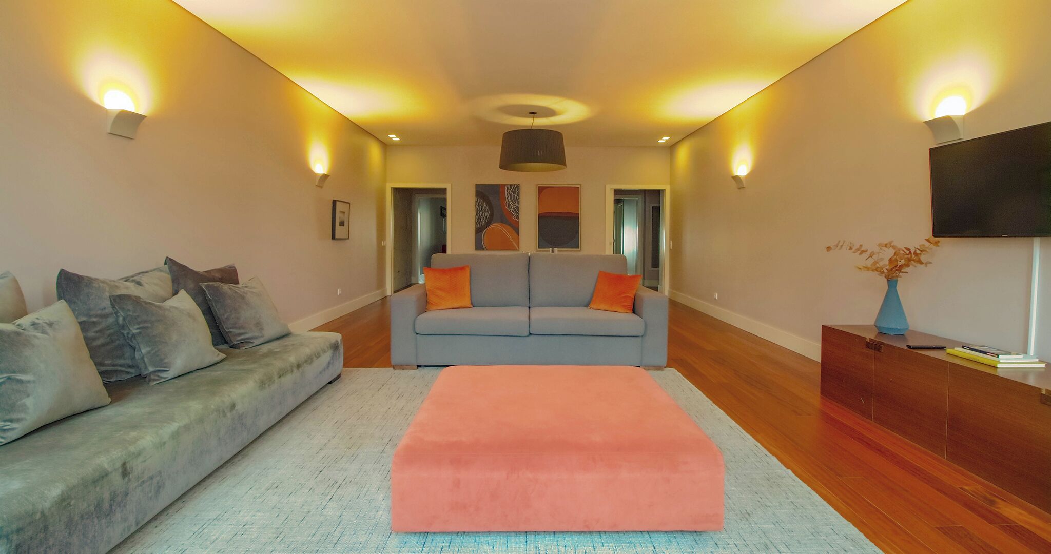 A modern living room with a grey sofa, a pink ottoman, and a blue sofa with orange cushions. There are wall-mounted lights and a flat-screen TV on a wooden cabinet.