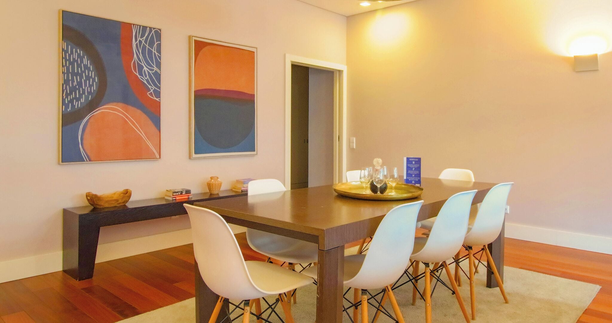A dining area with a wooden table, white chairs, and abstract paintings on the wall.