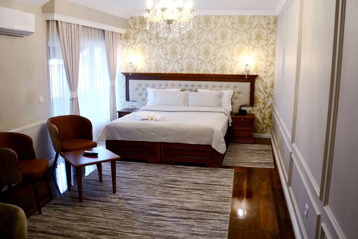 A hotel room with a large bed, a sitting area with a table and chairs, a chandelier, and patterned wallpaper.