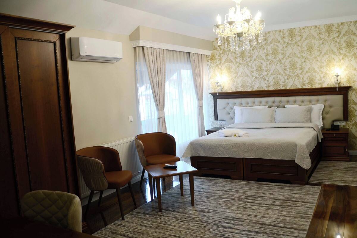 A hotel room with a large bed, a wooden wardrobe, a seating area with two chairs, a small table, and a chandelier.