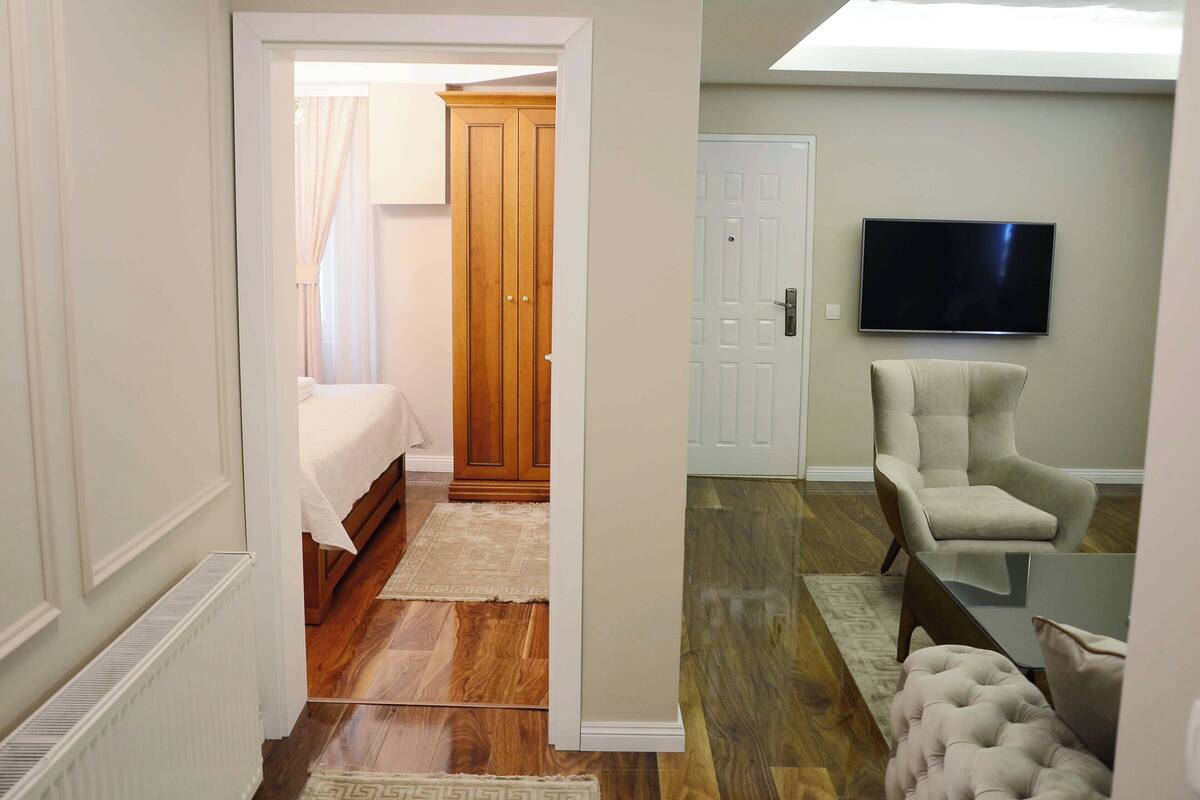A bedroom with a bed, a wardrobe, a television, and a seating area with a chair and ottoman.
