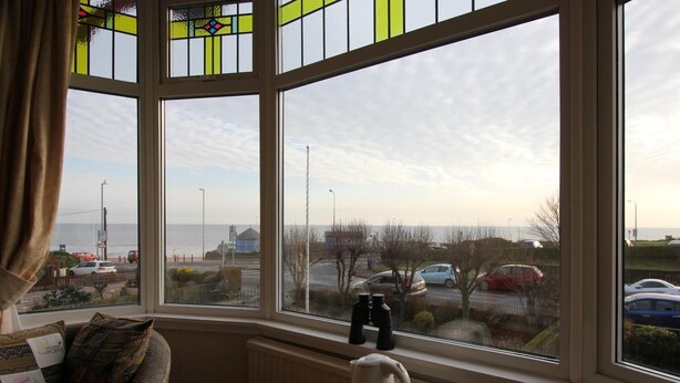 A room with a bay window overlooking a parking area and the sea.
