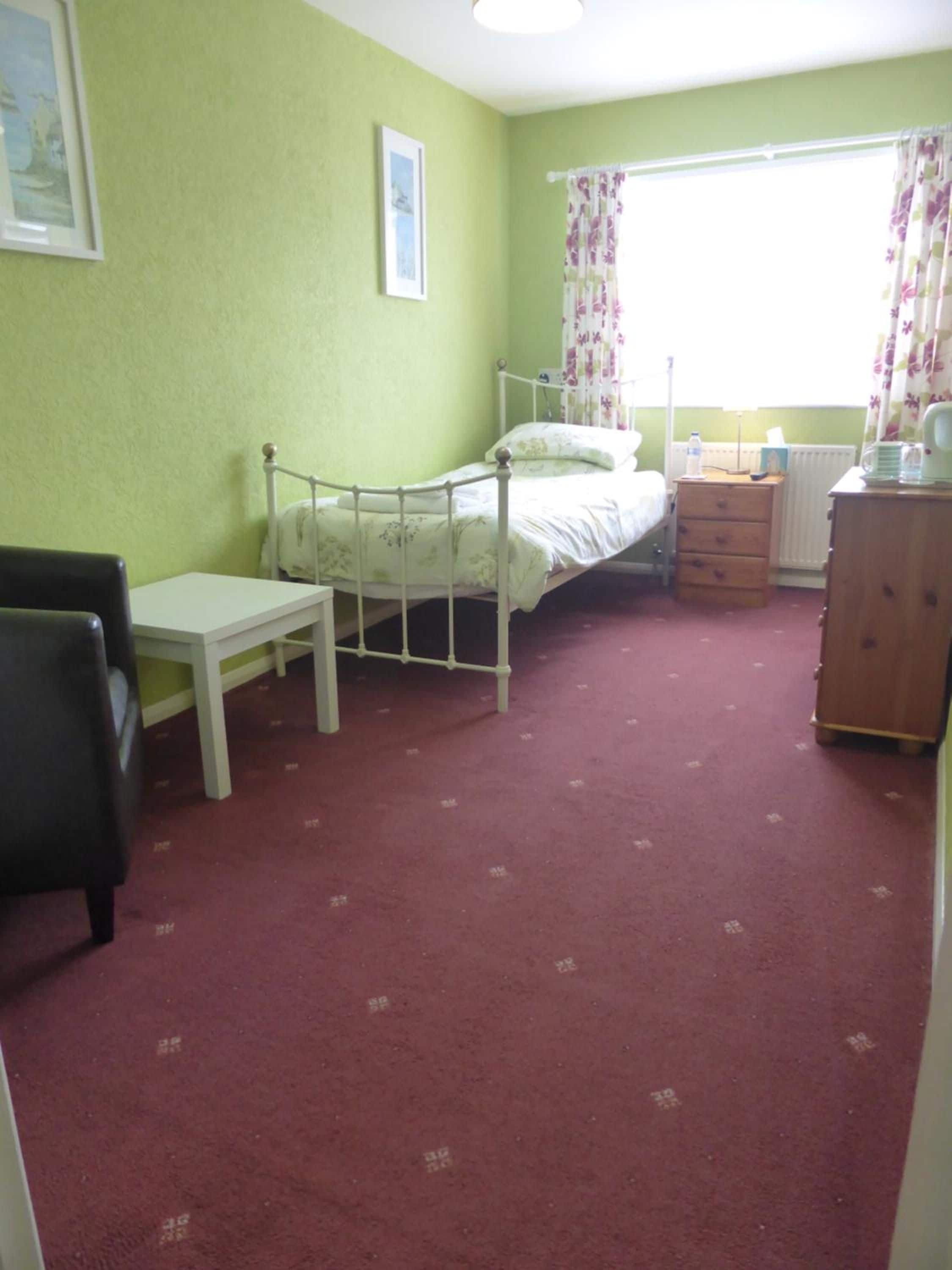A small, single-bed room with a wooden dresser, a bedside table, and a window with floral curtains.