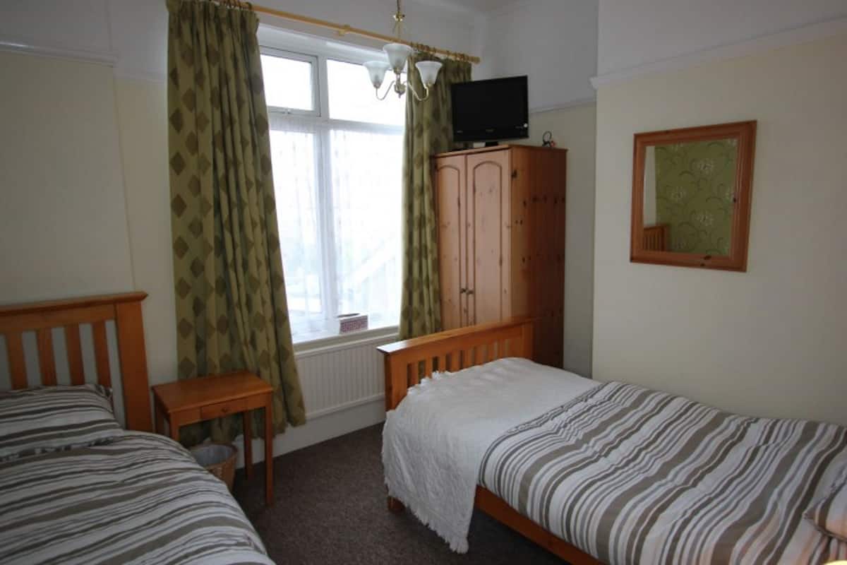 A bedroom with two single beds, a wooden wardrobe, a television on top, and a window with curtains.