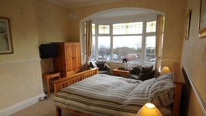A bedroom with a bed, a television, a wooden cabinet, a window with curtains, and a view of the outside.