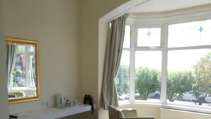 A bedroom with a bed, a window with curtains, a chair, a small table, and a shelf with items.