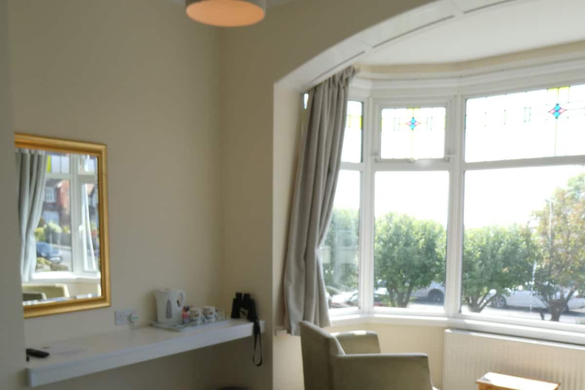 A bedroom with a bed, a window with curtains, a chair, a small table, and a shelf with items.