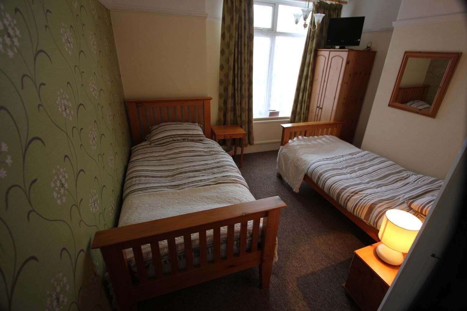 A bedroom with two single beds, a wooden wardrobe, a television on a cabinet, and a window with curtains.
