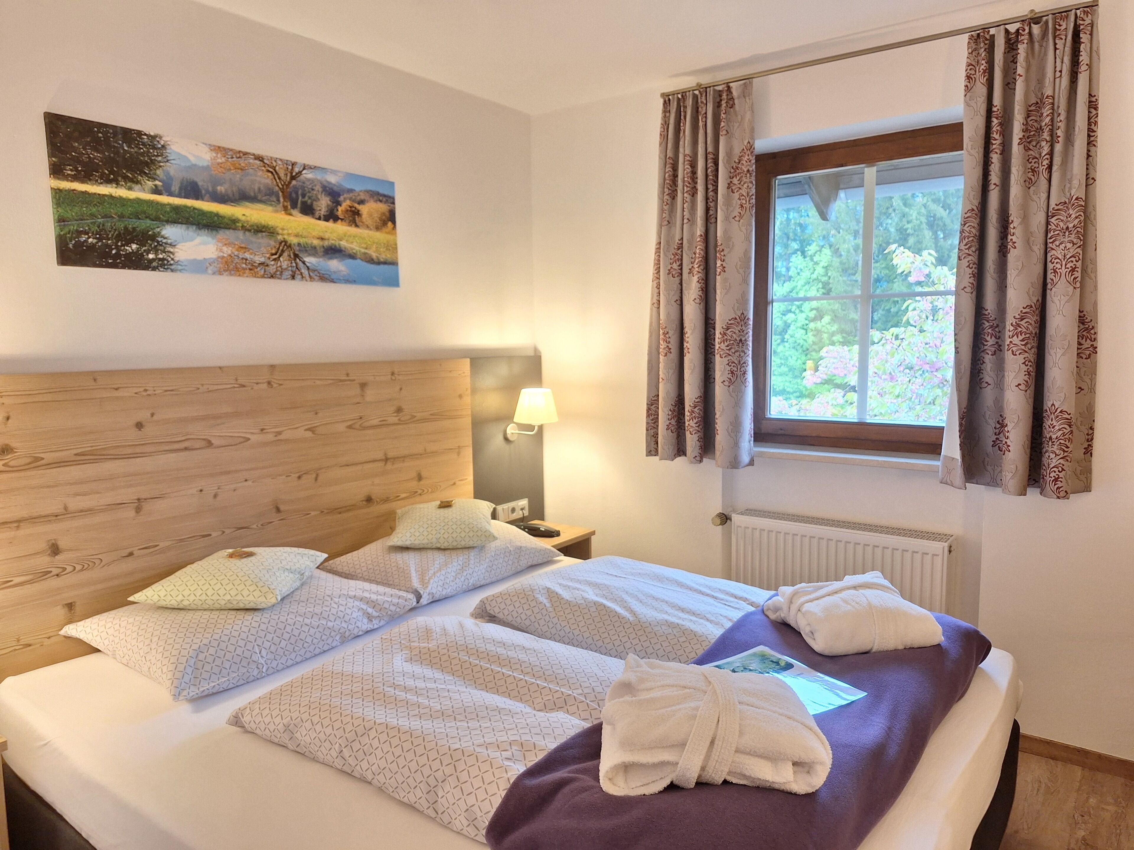 Ein ordentlich bezogenes Bett mit gemusterten Kissen, einer Nachttischlampe und einem Fenster mit Vorhängen.