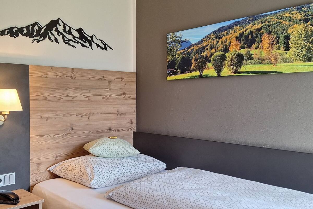 A hotel room with a bed, a nightstand, a lamp, a mountain wall decal, and a scenic painting.