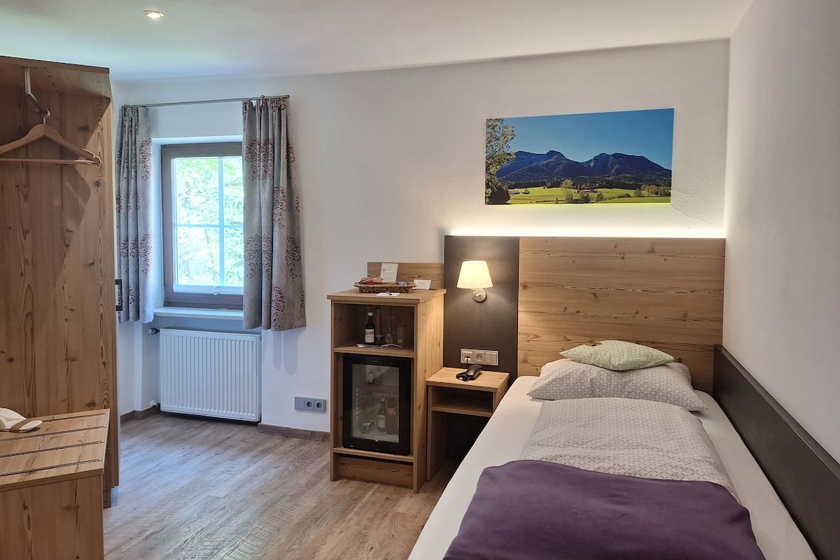 A hotel room with a bed, a nightstand, a wooden wardrobe, a window with curtains, a framed picture on the wall, and a radiator.