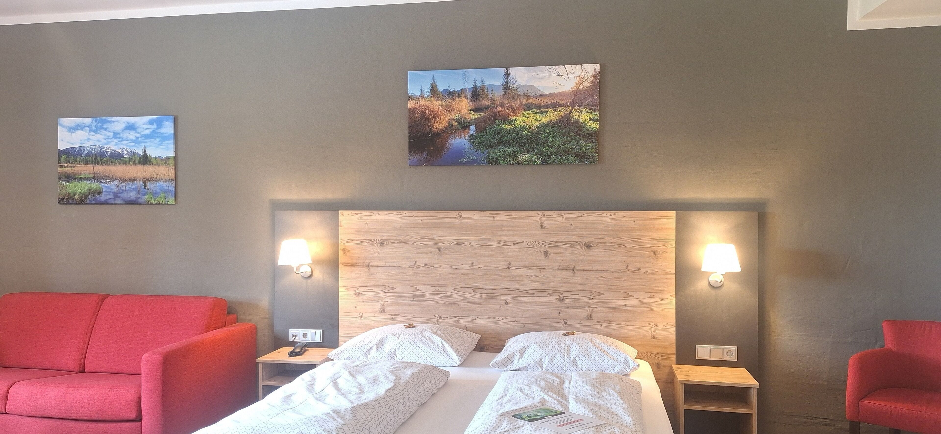 Una habitación de hotel con un cabecero de madera, dos mesitas de noche con lámparas, una cama con ropa blanca y dos sillones rojos.
