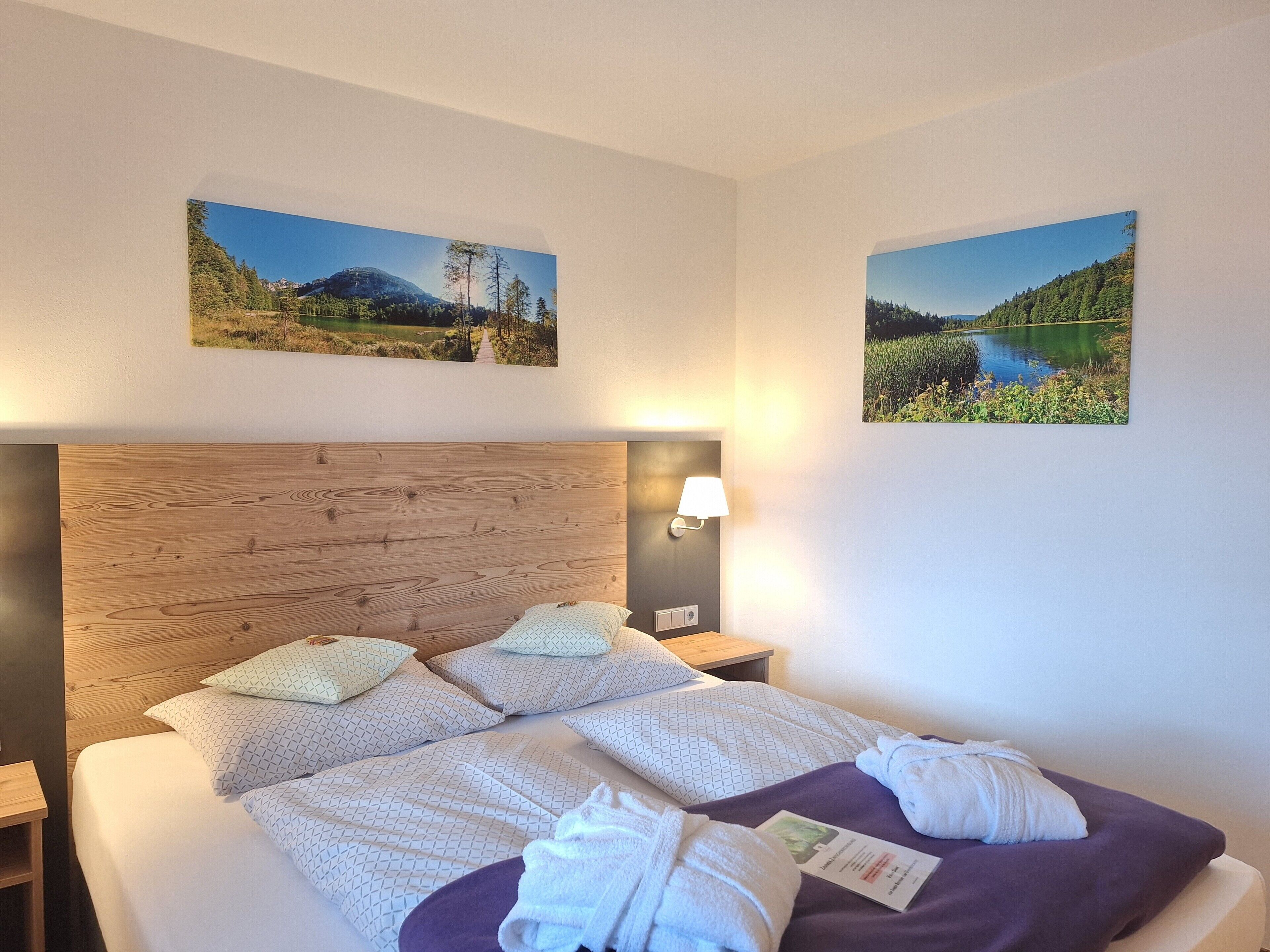 A hotel room with a wooden headboard, two beds with patterned bedding, a nightstand with a lamp, and two landscape paintings on the walls.