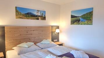 A hotel room with a wooden headboard, two beds with patterned bedding, a nightstand with a lamp, and two landscape paintings on the walls.