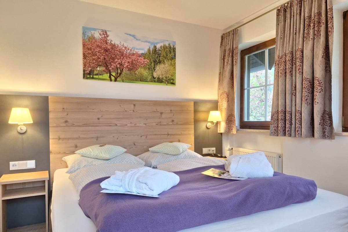 A hotel room with a wooden headboard, two bedside lamps, a bed with a purple blanket, and a window with patterned curtains.