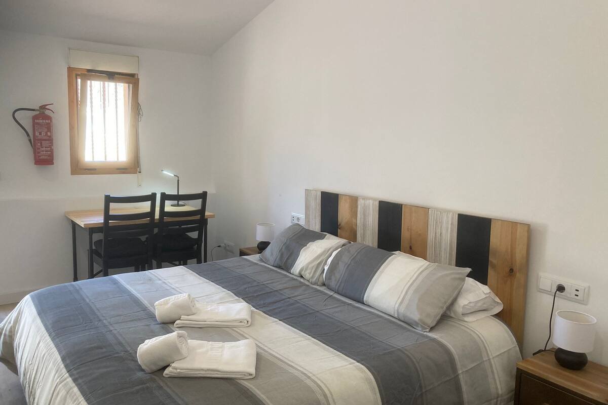 Ein Schlafzimmer mit Bett, Schreibtisch, zwei Stühlen, einem Fenster und einem Feuerlöscher.