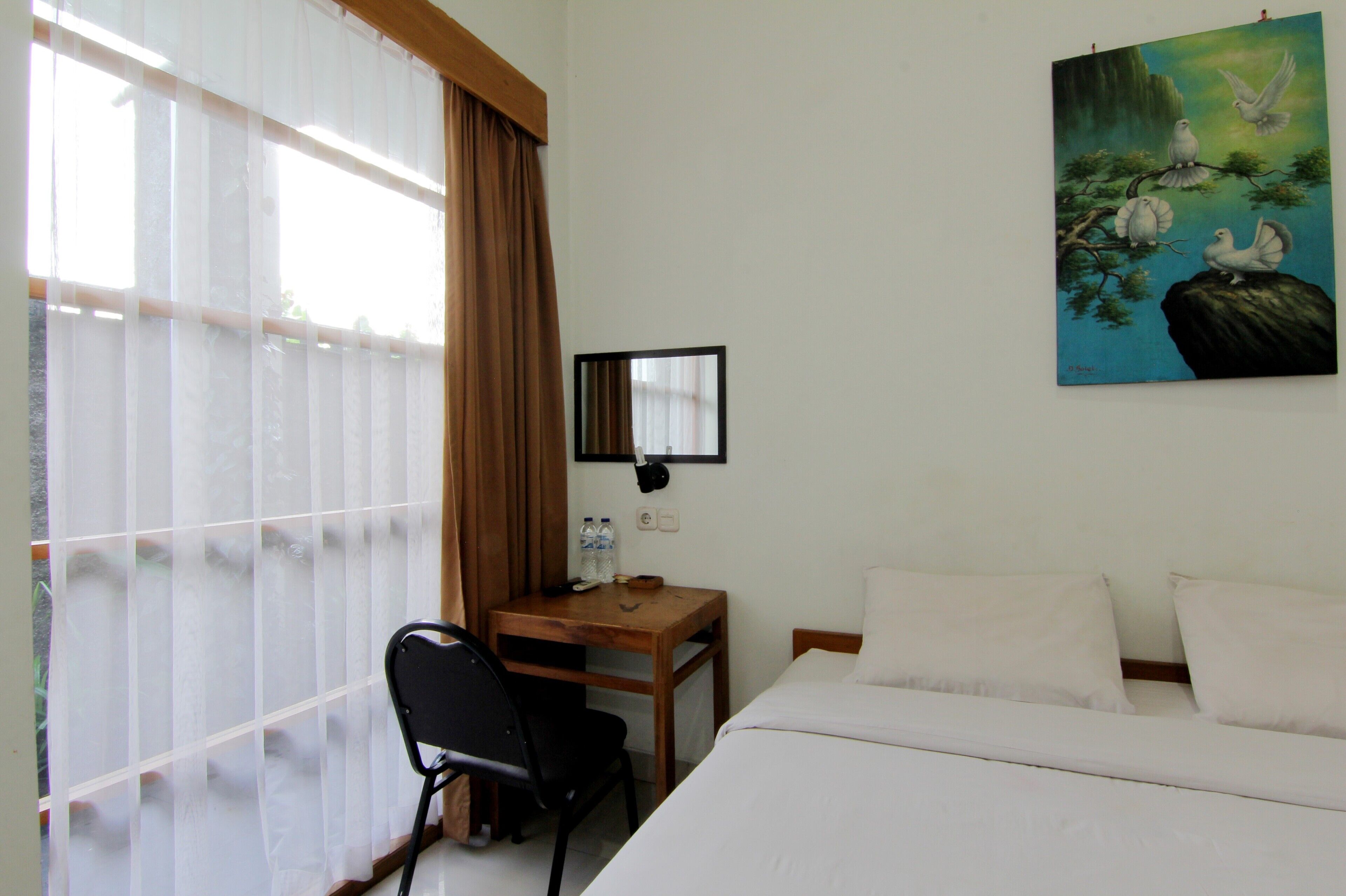 A hotel room with a bed, a desk, a chair, a window with curtains, and a painting on the wall.