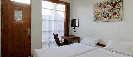 A hotel room with two beds, a wooden door, a window with curtains, a small table with a chair, and a wall-mounted TV.