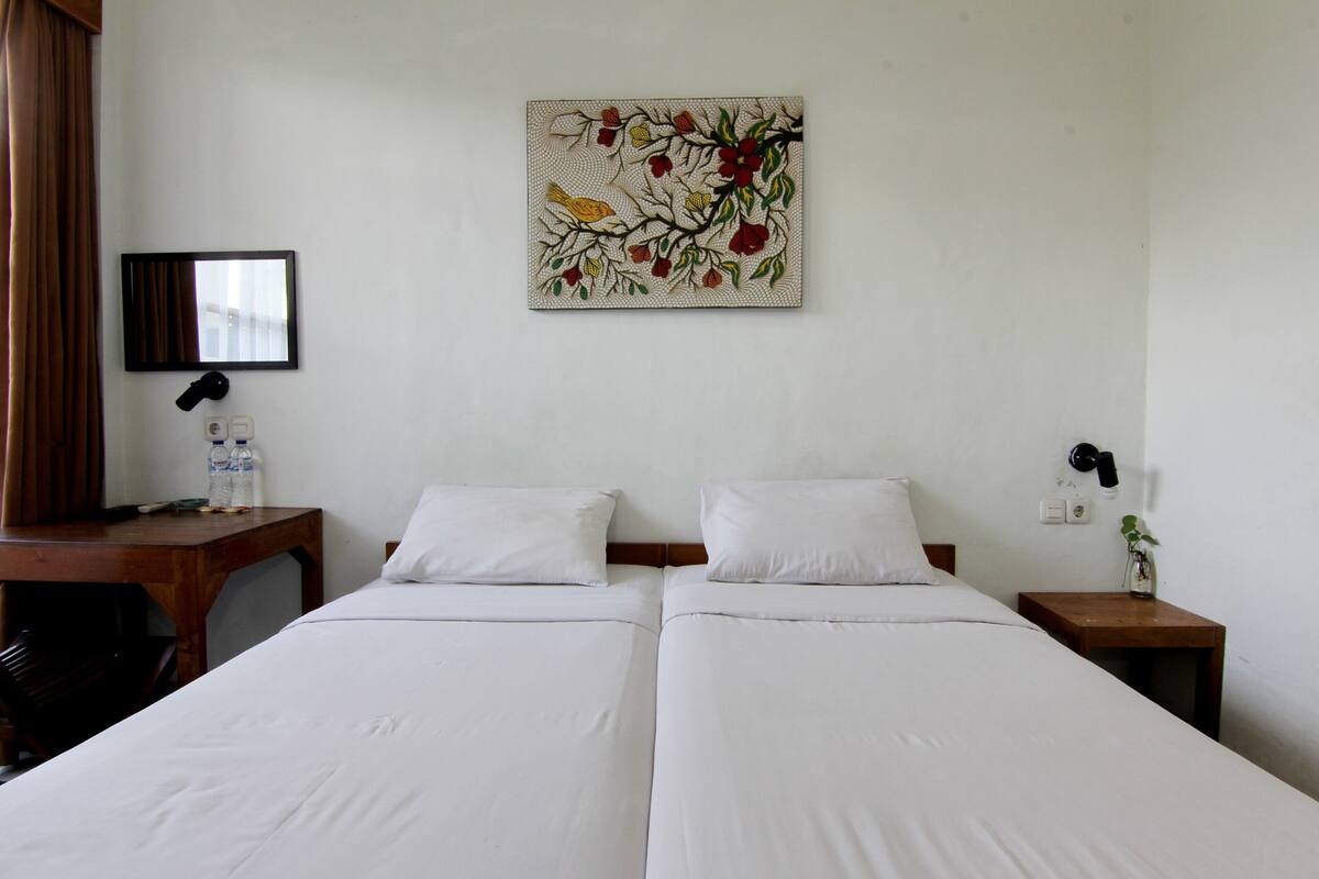 Une chambre d’hôtel avec deux lits simples, une table de chevet en bois avec une lampe, une petite table avec une bouteille et des verres, et un miroir fixé au mur.