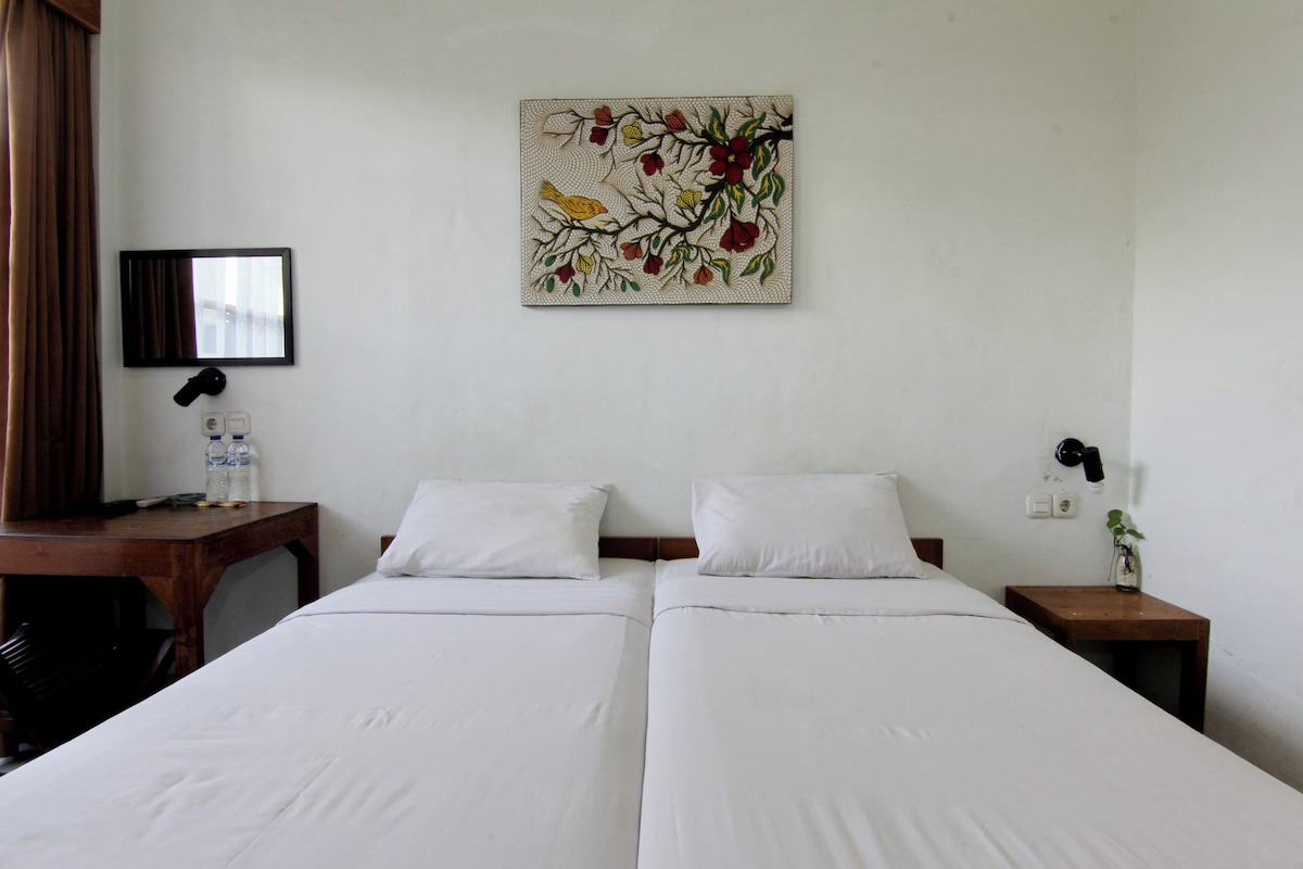 Habitación de hotel con dos camas individuales, un escritorio de madera, un espejo, una obra de arte en la pared y una mesita de noche con una planta.