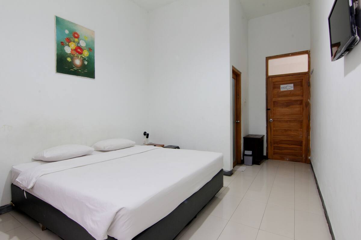 Ein Schlafzimmer mit einem Bett, einem Nachttisch, einem an der Wand befestigten Fernseher und einer Tür zu einem anderen Raum.