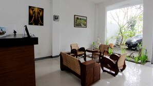Uma área de recepção com uma mesa de madeira, cadeiras de vime, uma mesa redonda e duas imagens emolduradas na parede.