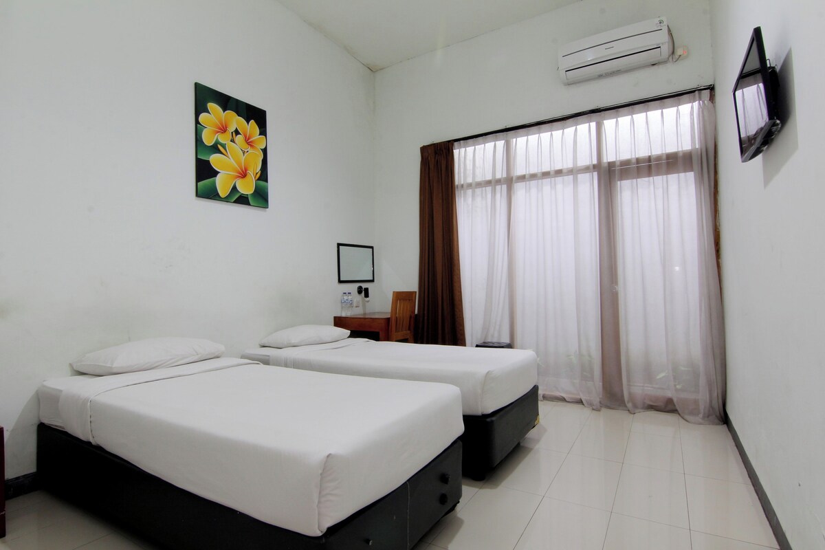 A hotel room with two beds, a TV, an air conditioner, a window with curtains, and a painting on the wall.