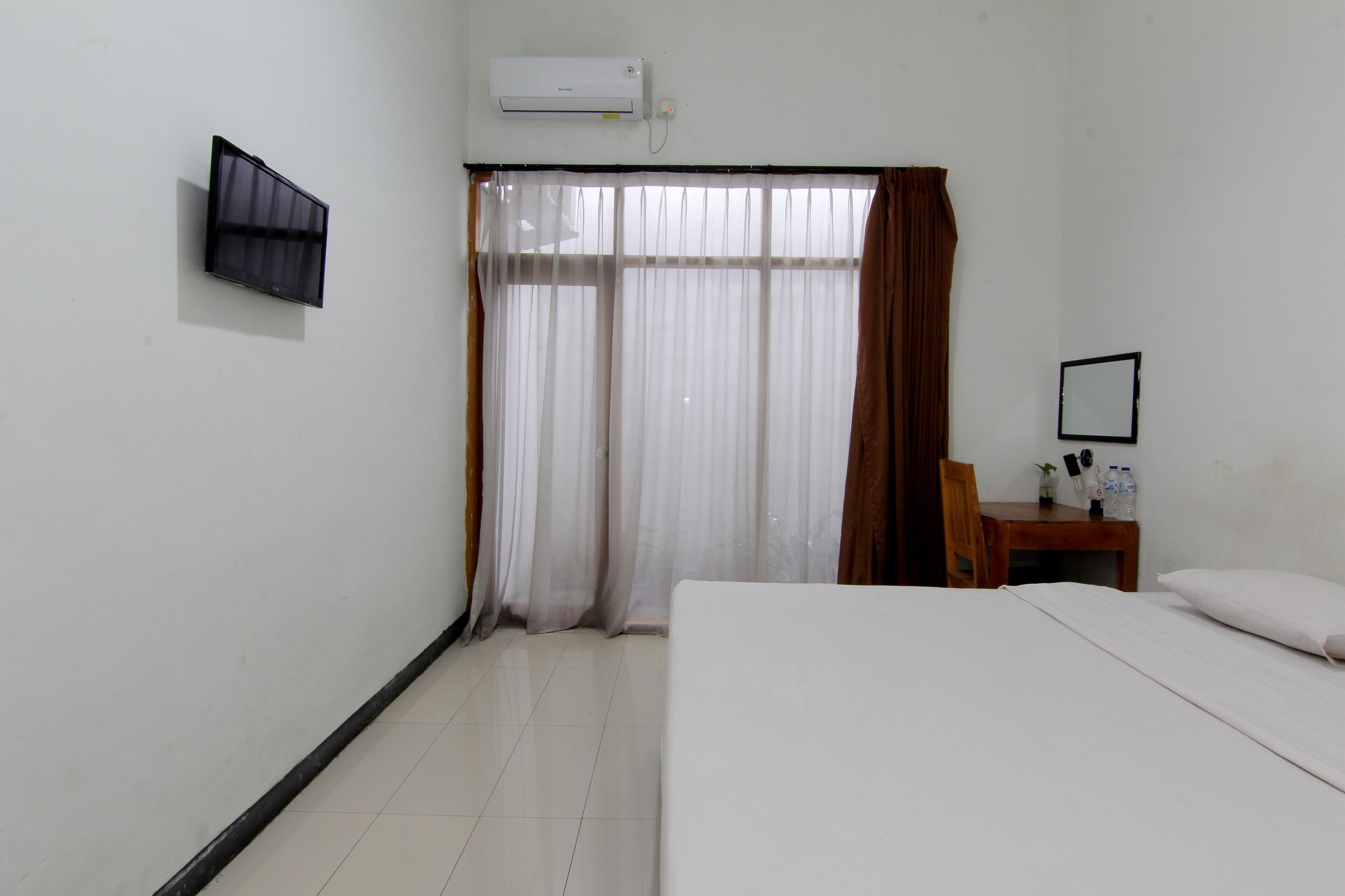 Habitación de hotel con cama, televisor montado en la pared, ventana con cortinas translúcidas y una pequeña mesa con una silla.