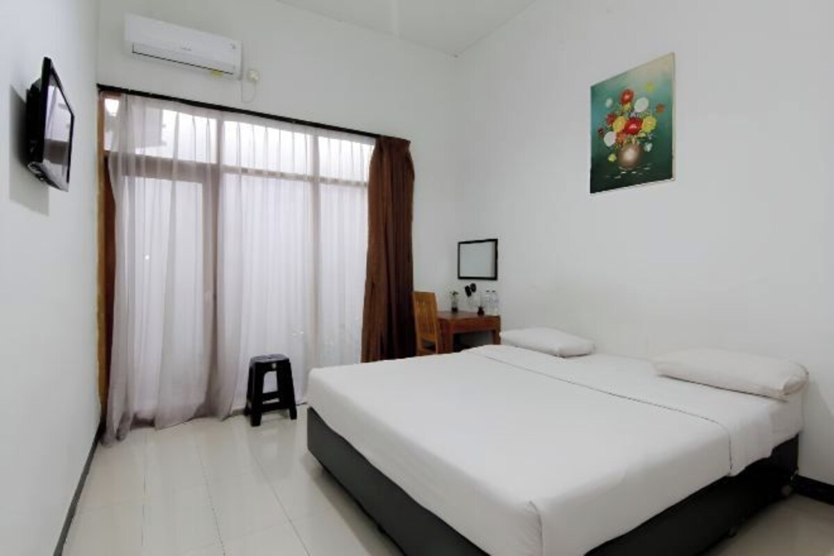 A hotel room with a bed, a desk, a chair, a small stool, and a painting on the wall.
