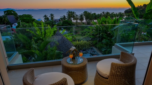 A balcony with wicker furniture and a glass table with drinks, overlooking a tropical garden and ocean view. - Bel Horizon (Nosy Be)