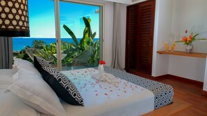 A bedroom with a bed, a wooden bench, a shelf with drinks, and a view of the ocean. - Bel Horizon (Nosy Be)