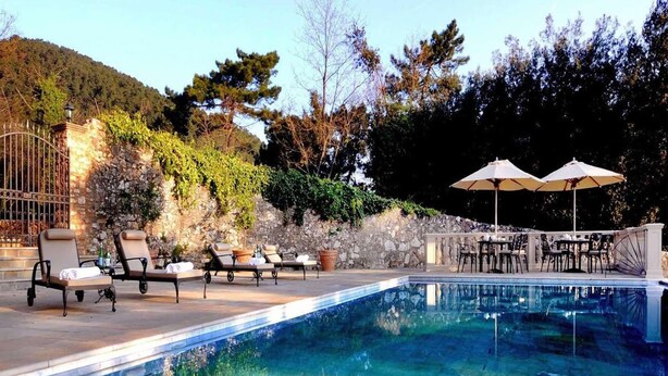 Zona de piscina con sillas de descanso y sombrillas, rodeada de muros de piedra y árboles.