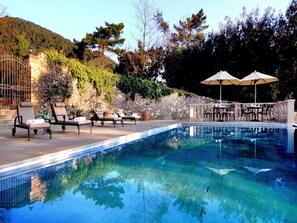 Zona de piscina con sillas de descanso y sombrillas, rodeada de muros de piedra y árboles.