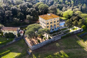Vista aerea di un ampio edificio a più piani con tetto in tegole, circondato da vegetazione rigogliosa e un muro in pietra.