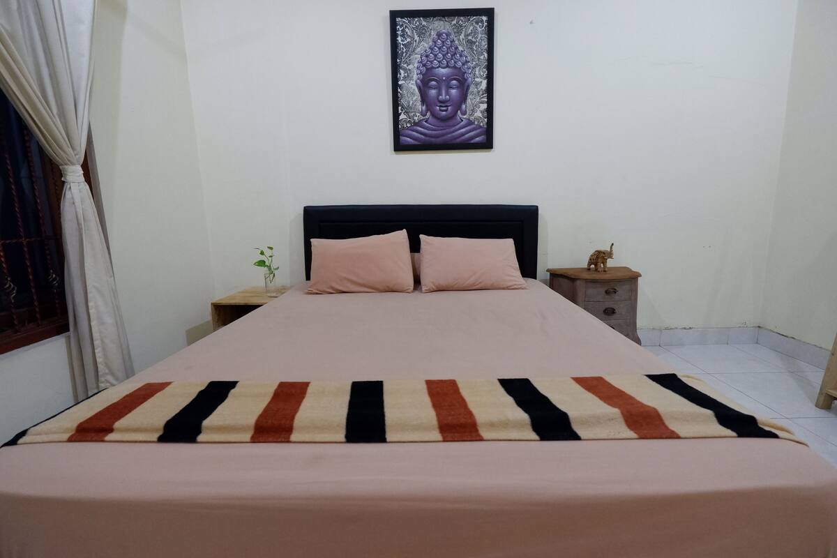 A neatly made bed with a striped blanket, a headboard, a wall-mounted Buddha artwork, and a small elephant figurine on a nightstand.