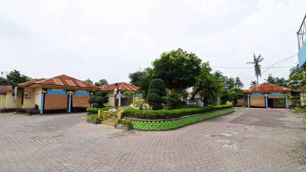 Un patio con suelo de baldosas, un área ajardinada central con un árbol, y edificios con puertas azules y techos rojos.