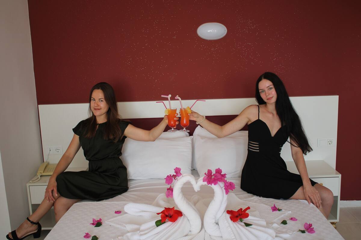 Two women sitting on a bed with a heart-shaped towel arrangement and drinks.