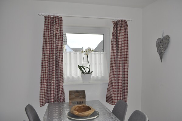Un comedor con una mesa, sillas, una planta en maceta, un letrero de madera que dice “LETTERS” y una ventana con cortinas cuadriculadas.