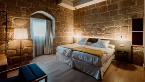 Una camera da letto con parete in pietra, un letto con lenzuola bianche, un comodino, una lampada e una finestra con tende.