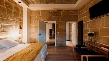 A bedroom with a stone wall, a bed with a wooden headboard, a bench, and a TV mounted on the wall.