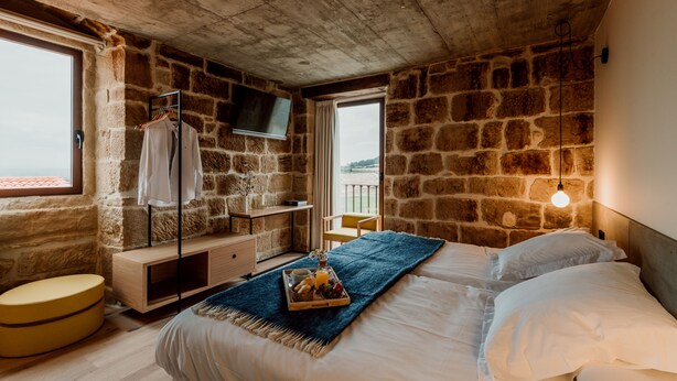 Camera da letto con pareti in pietra, un letto con lenzuola bianche, un comodino in legno, un televisore montato a parete e una finestra con vista.