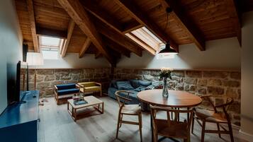 A cozy loft-style room with a stone wall, wooden ceiling beams, a round wooden table, and a blue sofa.